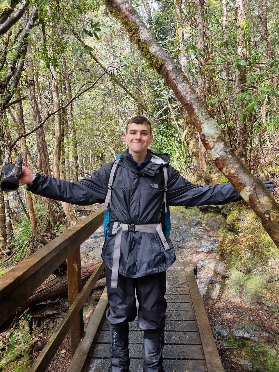 Man in black rain jacket and hiking gear smiling with arms outstretched on a forest trail wooden bridge.