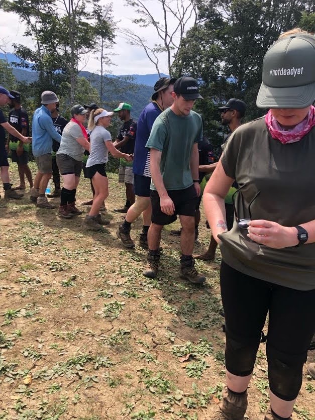 Group of hikers on a trail outdoors, shaking hands in a line with trees and mountains in the background; one person in front wearing a green cap with #notdeadyet.