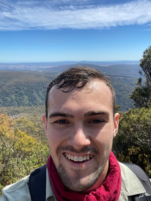 Smiling man with short dark hair and light beard wearing a red scarf and backpack, with a mountainous forest landscape and blue sky in the background.