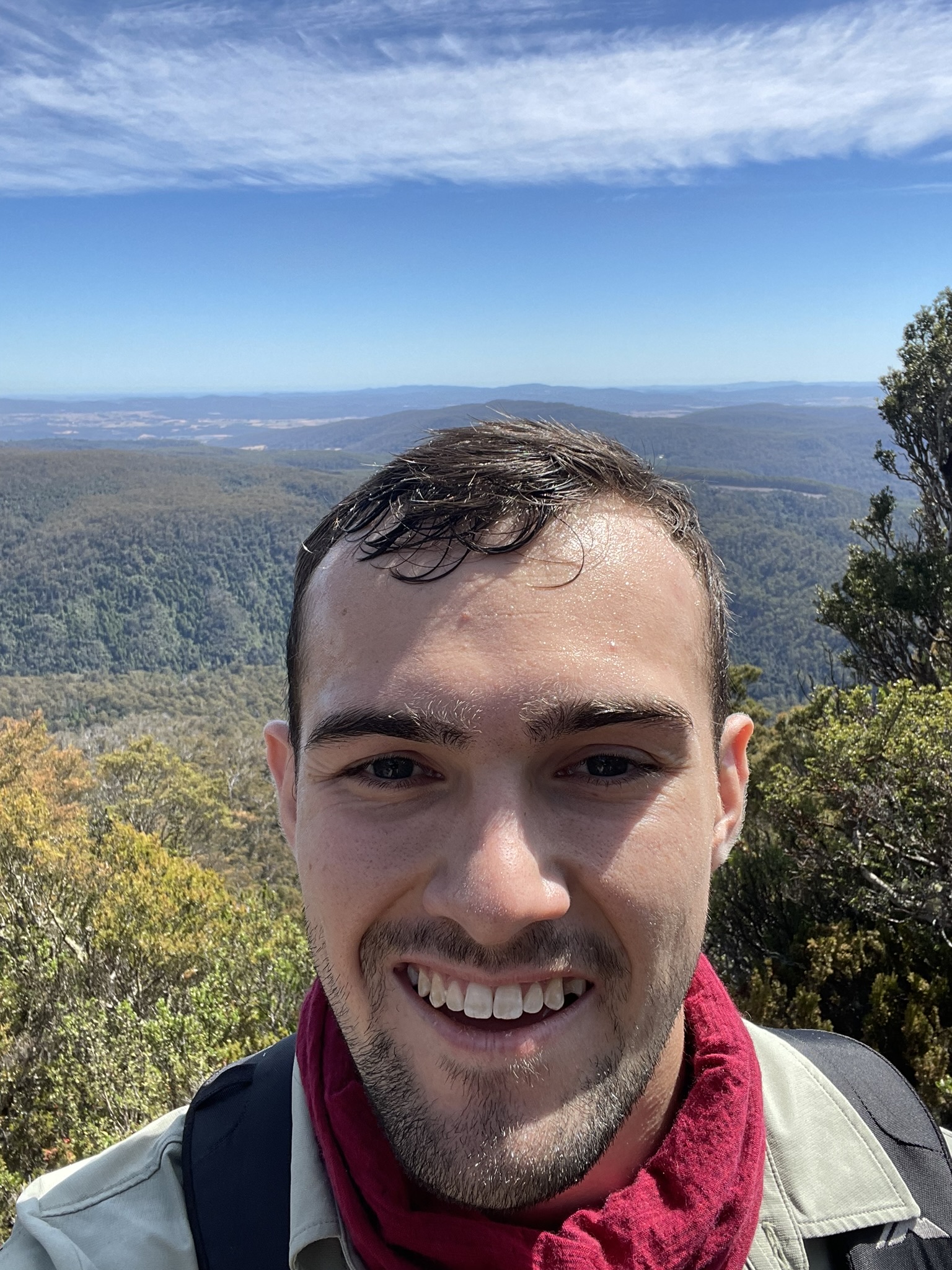 Smiling man with short dark hair and light beard wearing a red scarf and backpack, with a mountainous forest landscape and blue sky in the background.