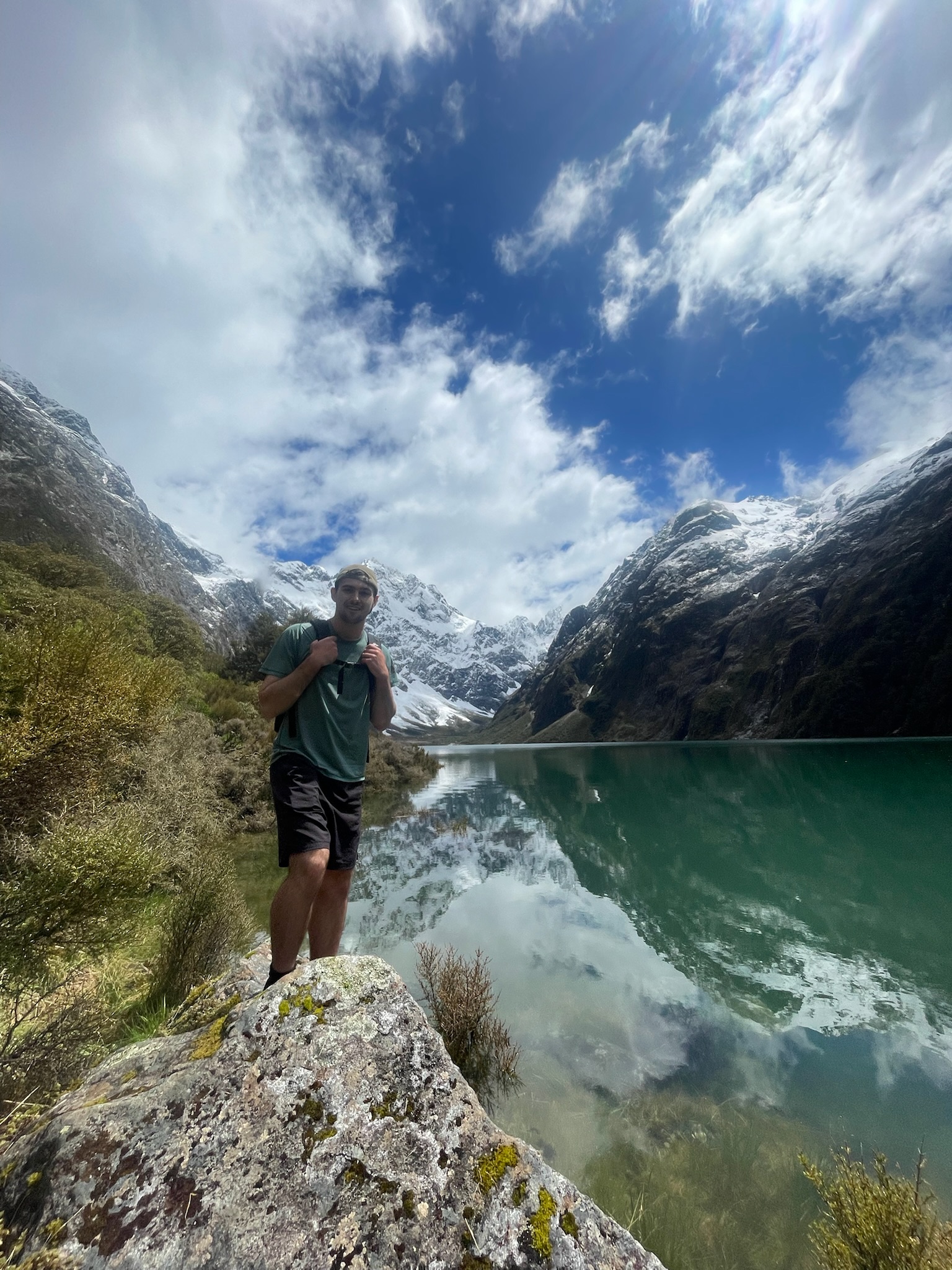 Man standing on a moss-covered rock beside a mountain lake with snow-capped peaks and a partly cloudy sky reflected in the water.