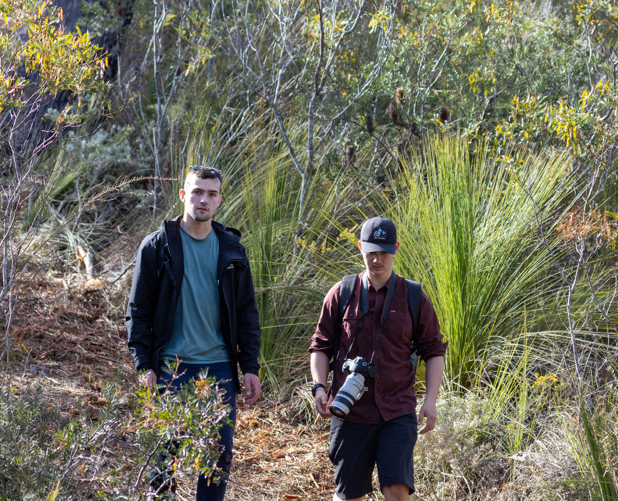 Two young men walking on a forest trail surrounded by dense green foliage, one carrying a camera with a large lens.