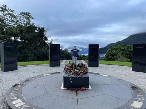 Person standing with arms open inside a circular memorial area with black pillars labeled Courage, Endurance, Mateship, and Sacrifice.