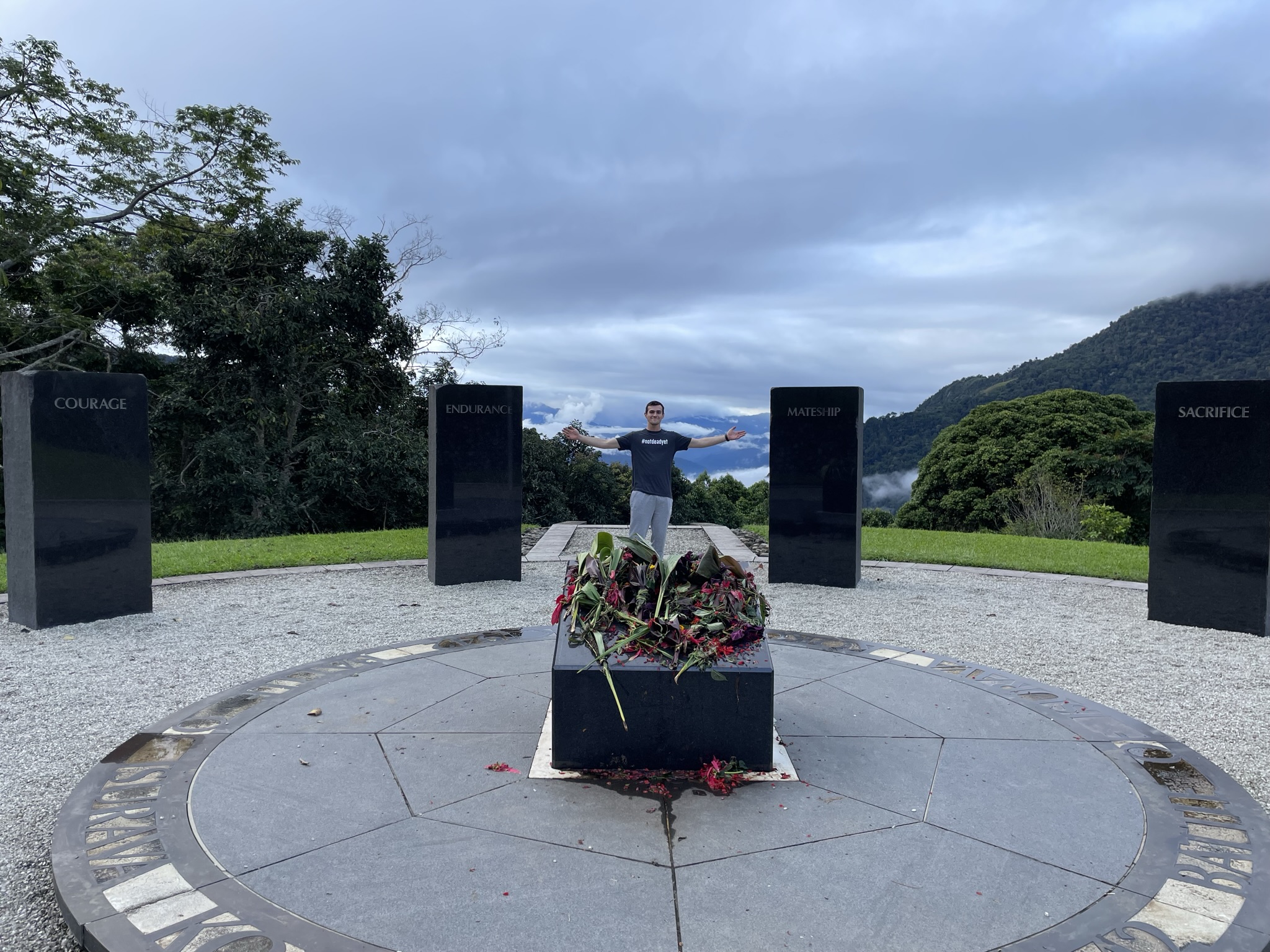 Person standing with arms open inside a circular memorial area with black pillars labeled Courage, Endurance, Mateship, and Sacrifice.