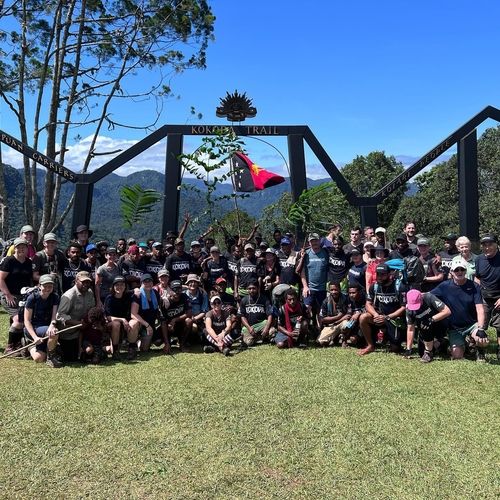 Aerial view of a single-story house with solar panels on the roof, surrounded by green lawn, dirt paths, scattered buildings, and dense forest extending to distant hills under a clear sky.