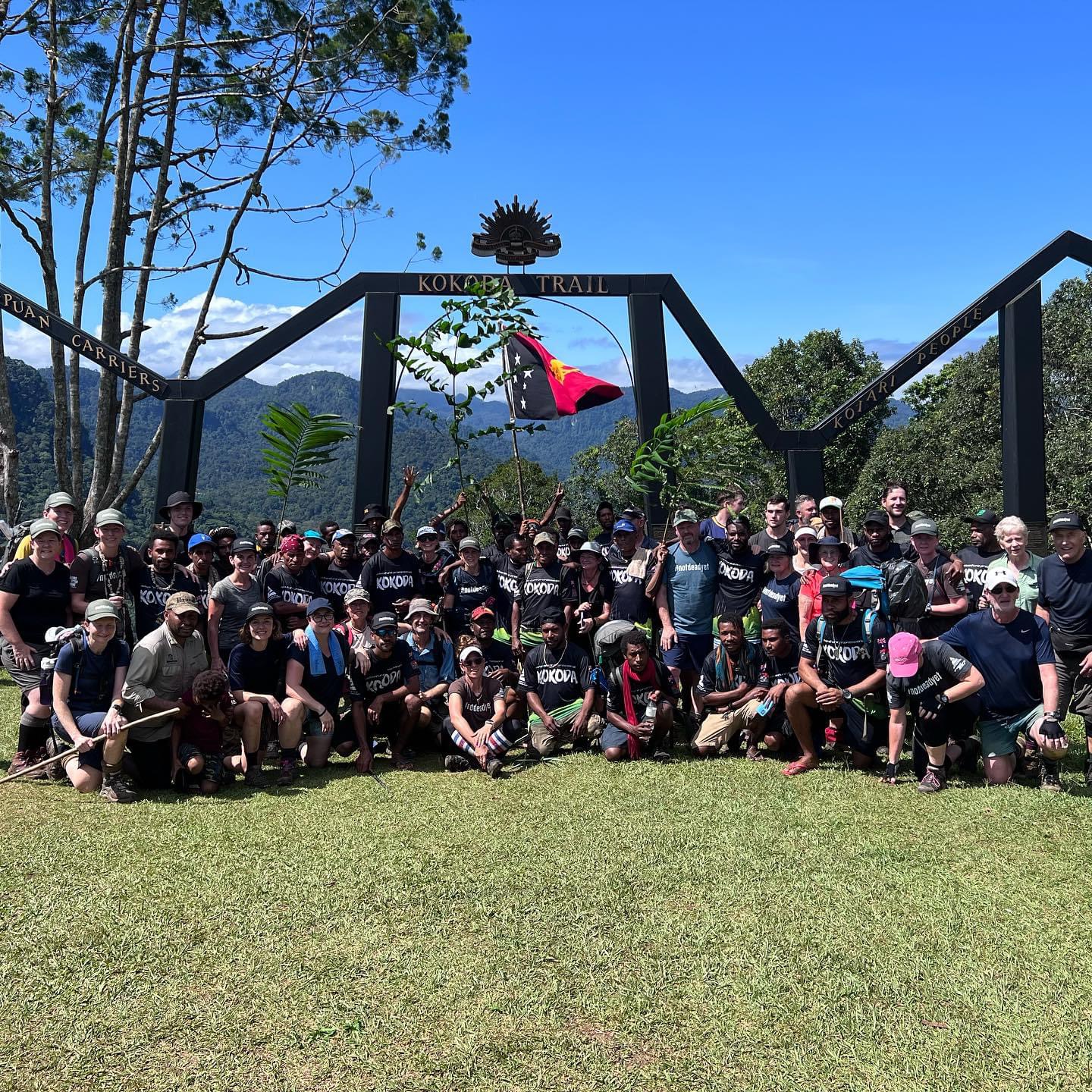 Aerial view of a single-story house with solar panels on the roof, surrounded by green lawn, dirt paths, scattered buildings, and dense forest extending to distant hills under a clear sky.
