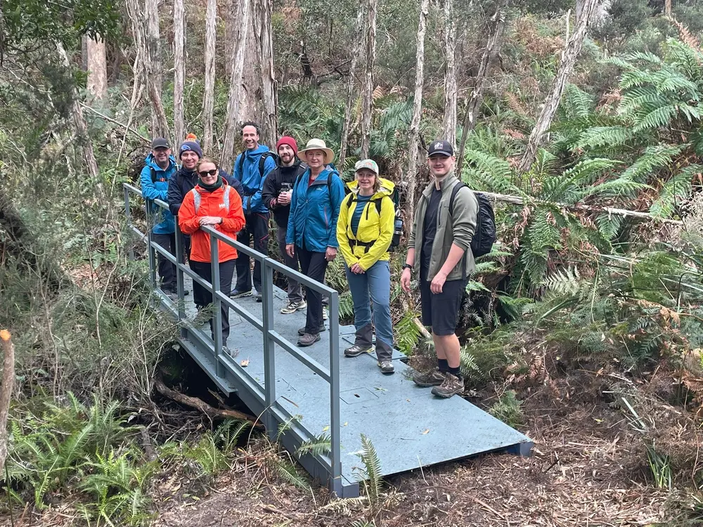 Group of eight hikers standing on a metal bridge in a dense forest with tall trees and ferns.