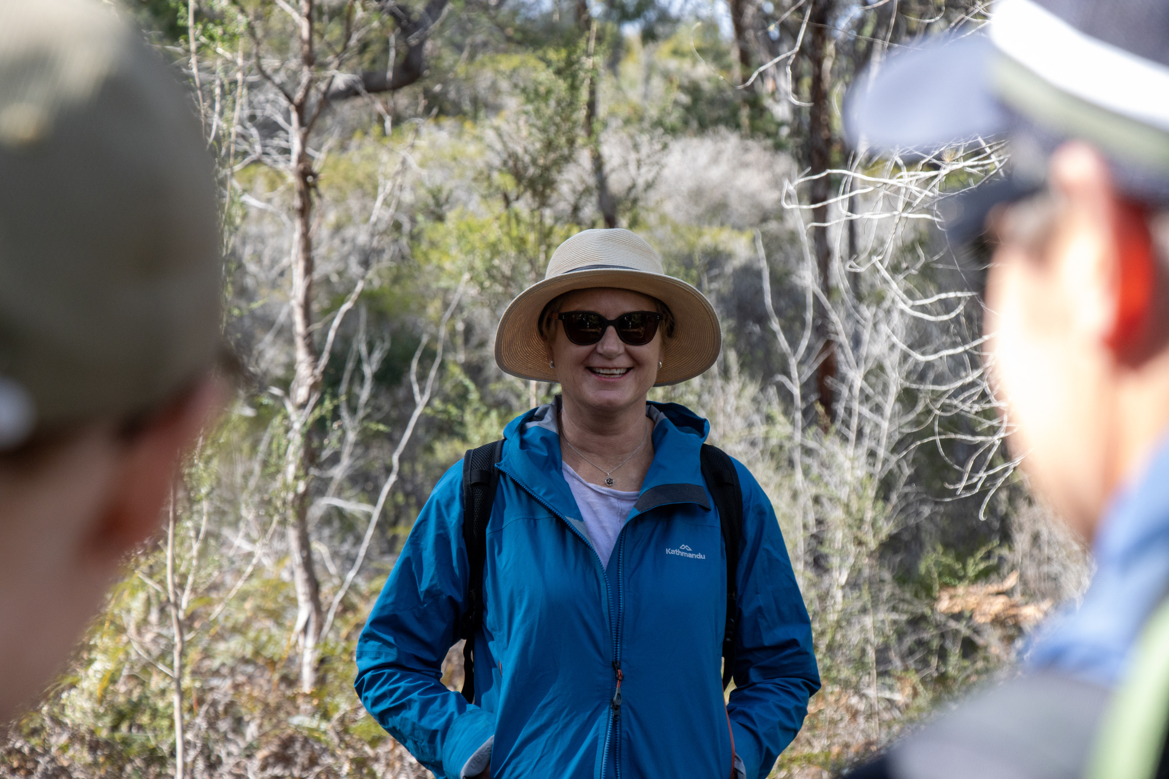 Aerial view of a single-story house with solar panels on the roof, surrounded by green lawn, dirt paths, scattered buildings, and dense forest extending to distant hills under a clear sky.