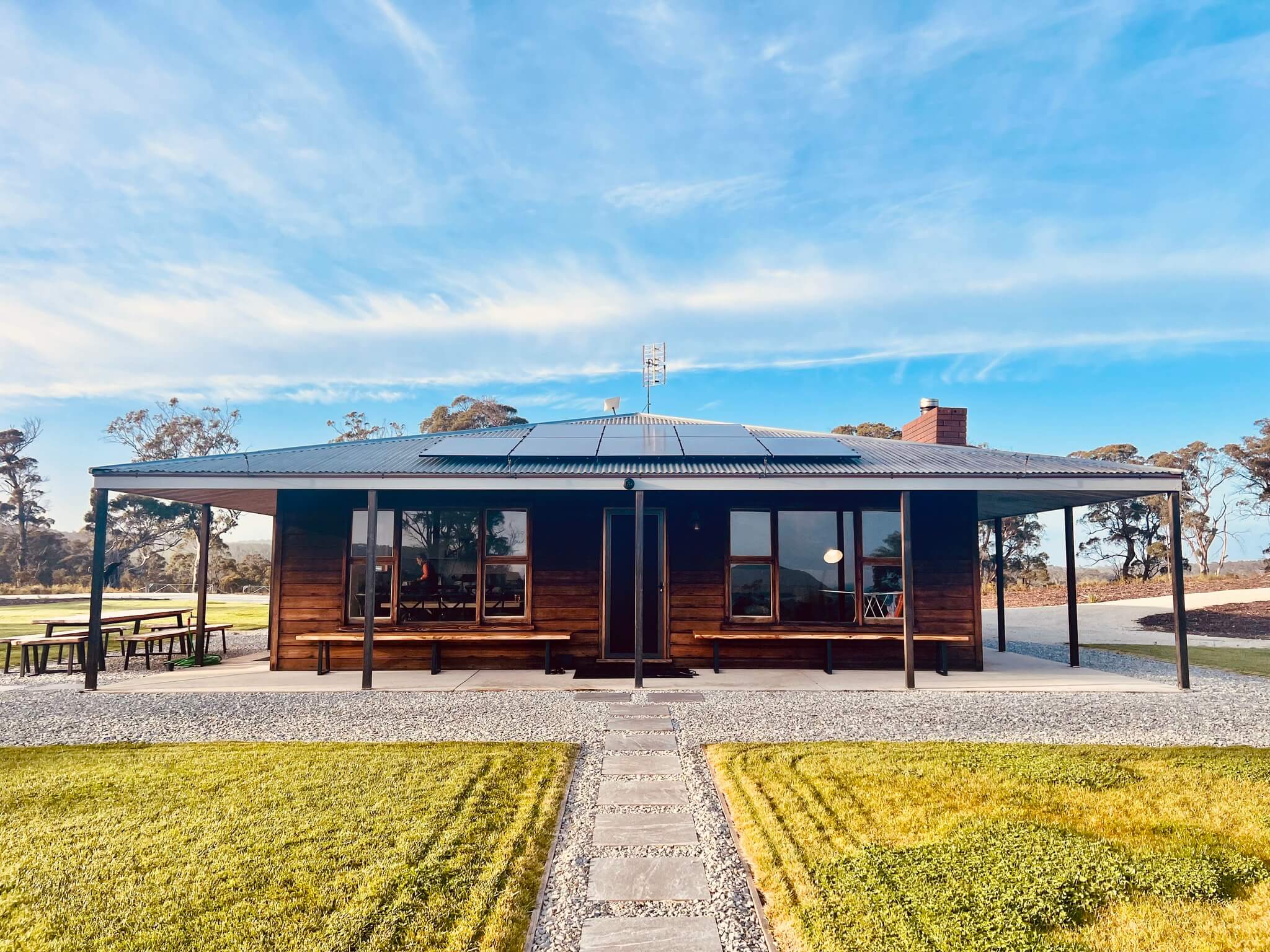 Modern wooden house with a metal roof equipped with solar panels, surrounded by green grass and under a blue sky.