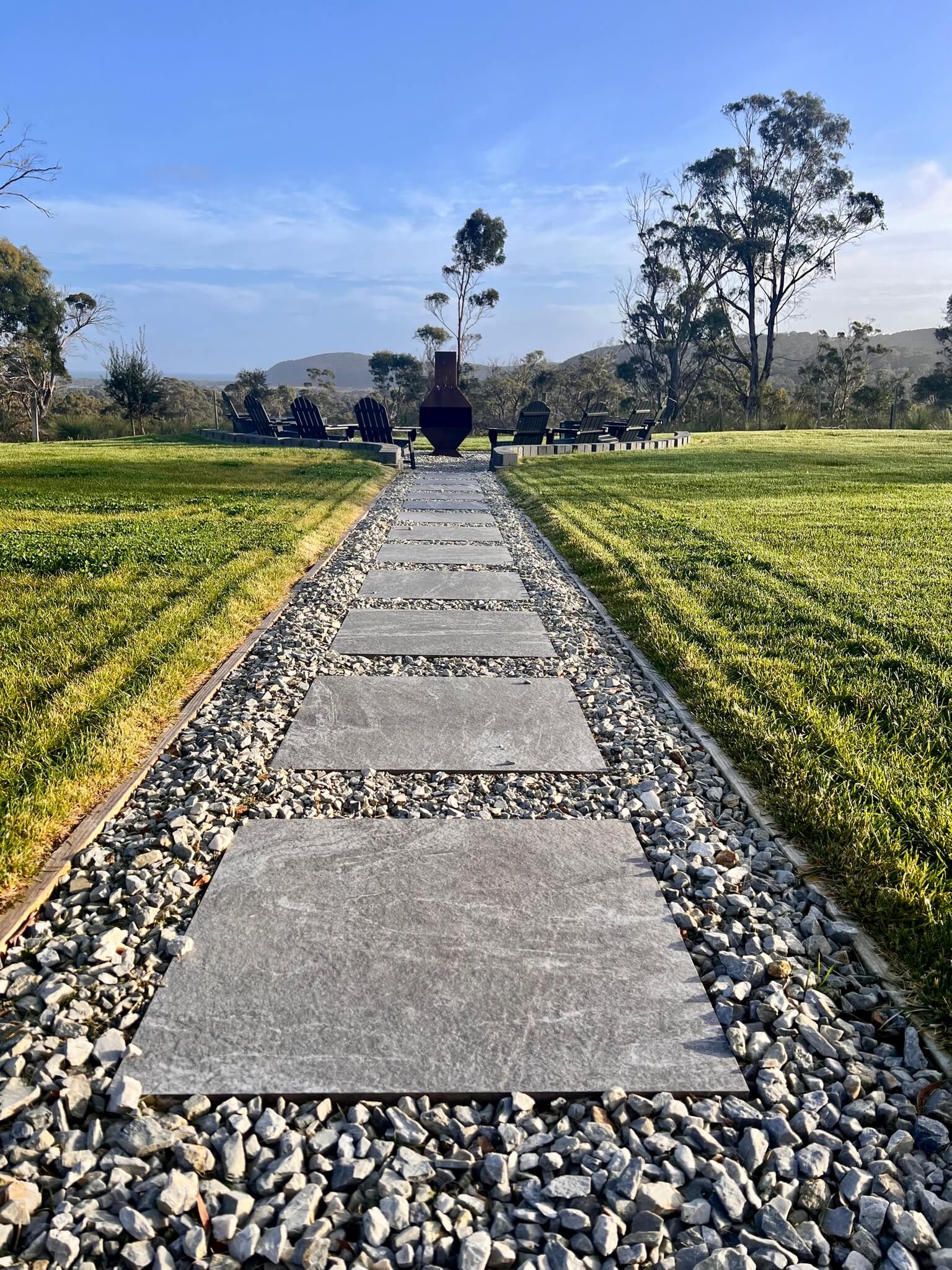 Outdoor setting with two white bathtubs and a wooden barrel sauna surrounded by trees and grass at sunset.