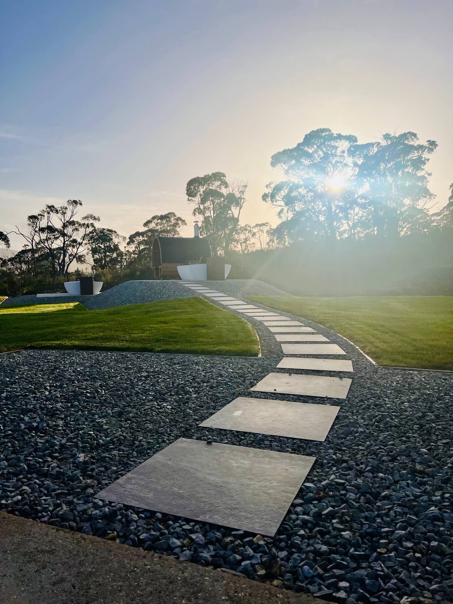 Stone pathway through gravel and grass leading to a small wooden structure with a curved roof, under a bright sun and surrounded by trees.