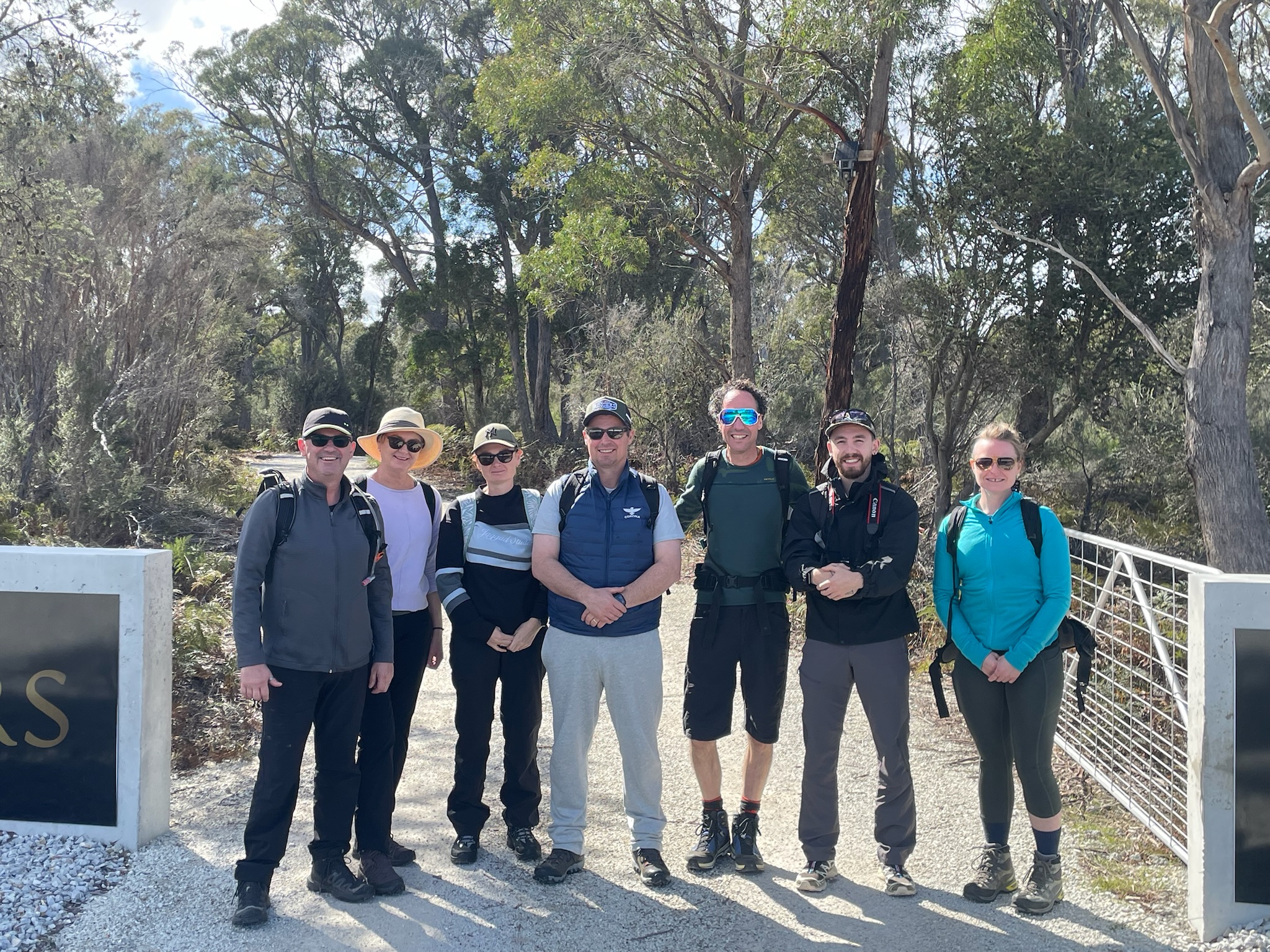Aerial view of a single-story house with solar panels on the roof, surrounded by green lawn, dirt paths, scattered buildings, and dense forest extending to distant hills under a clear sky.
