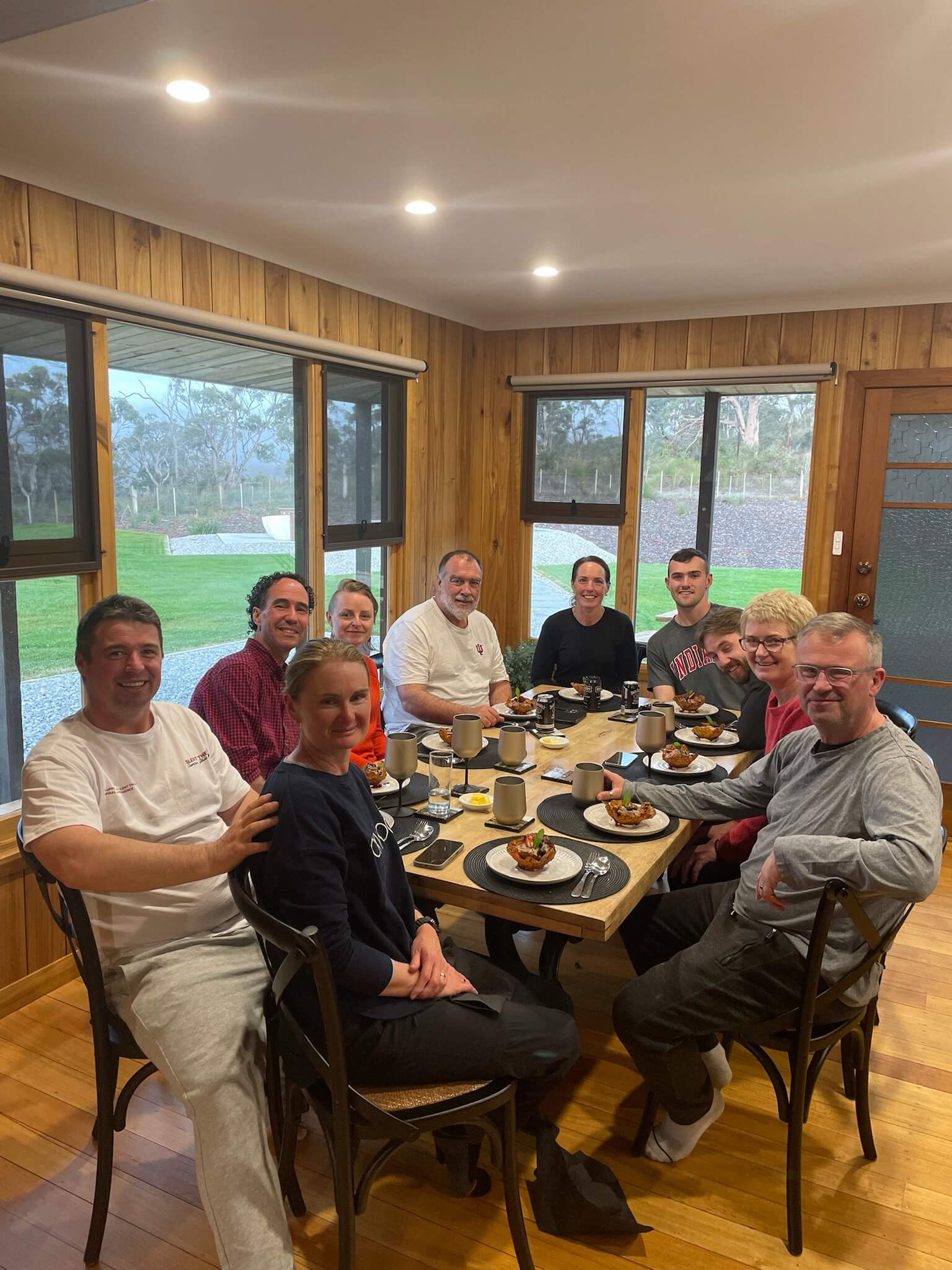 Group of ten people sitting around a wooden dining table with dessert plates and drinks, inside a room with wood-paneled walls and large windows showing a green outdoor view.