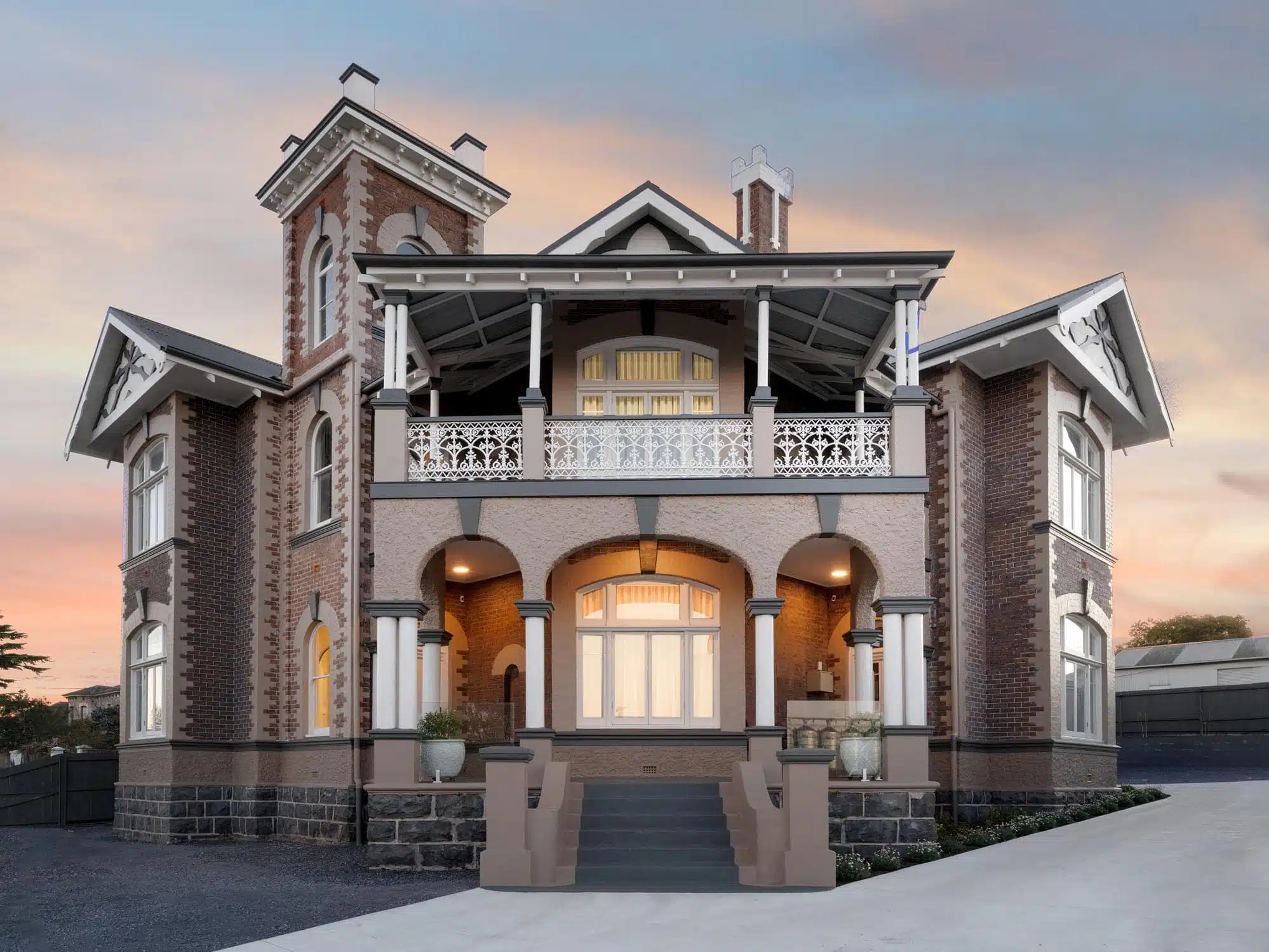 Colonial-style two-story house with brick and stone exterior, a front porch with arches, and a decorative iron balcony railing at sunset.