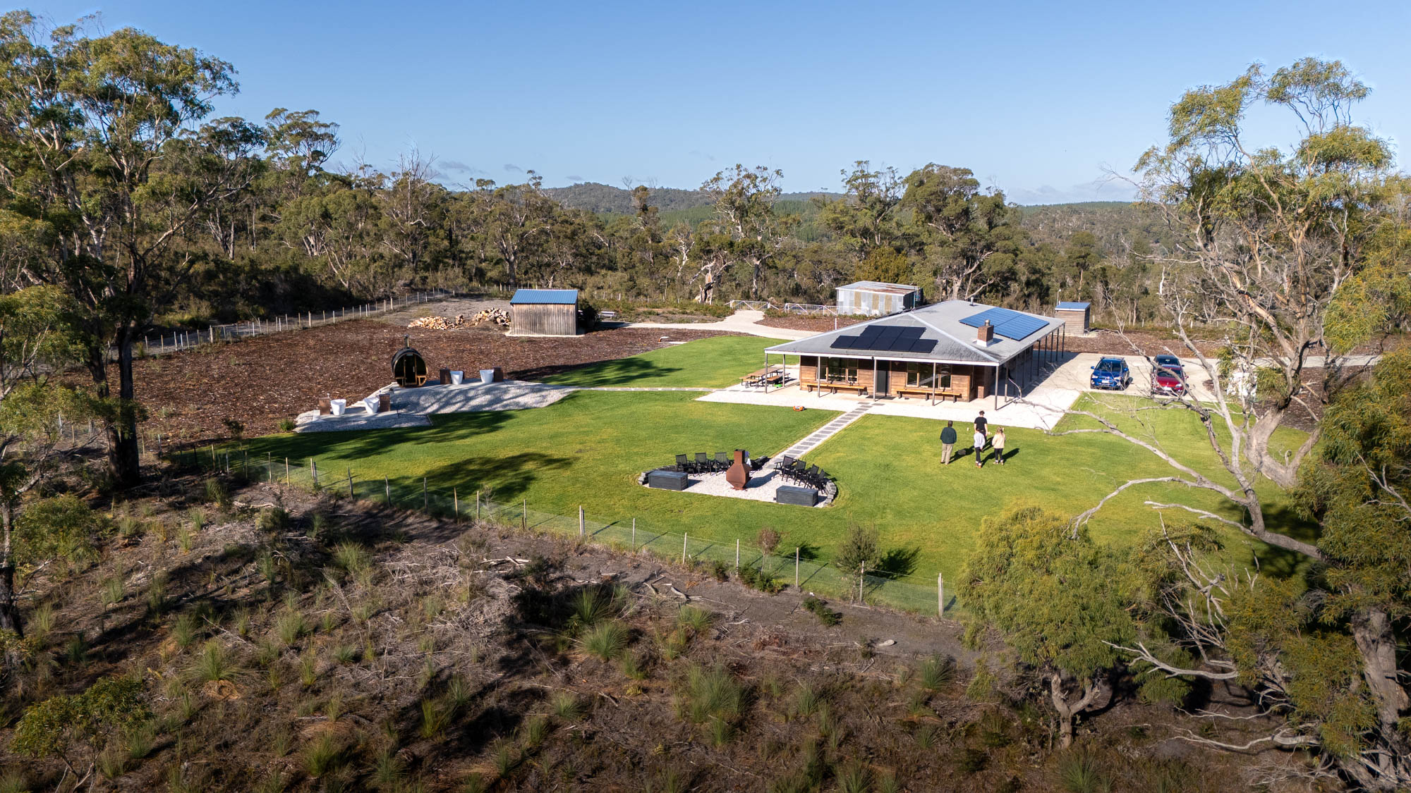 A group of people standing on a large green lawn near a wooden cabin with outdoor seating, surrounded by trees and nature.