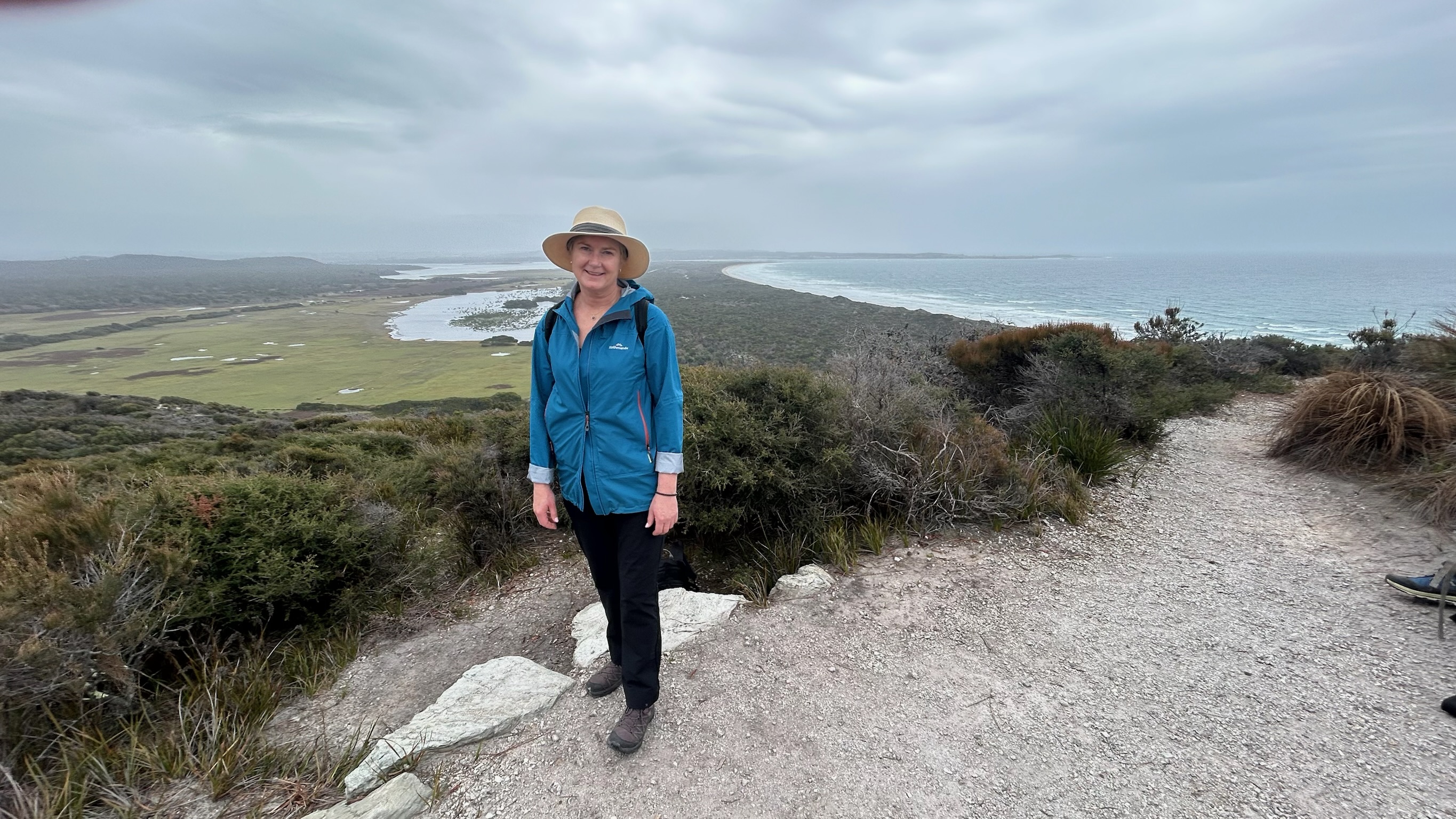 Woman in a blue jacket and beige hat standing on a hiking trail with coastal scenery of bushes, green fields, and ocean in the background under a cloudy sky.