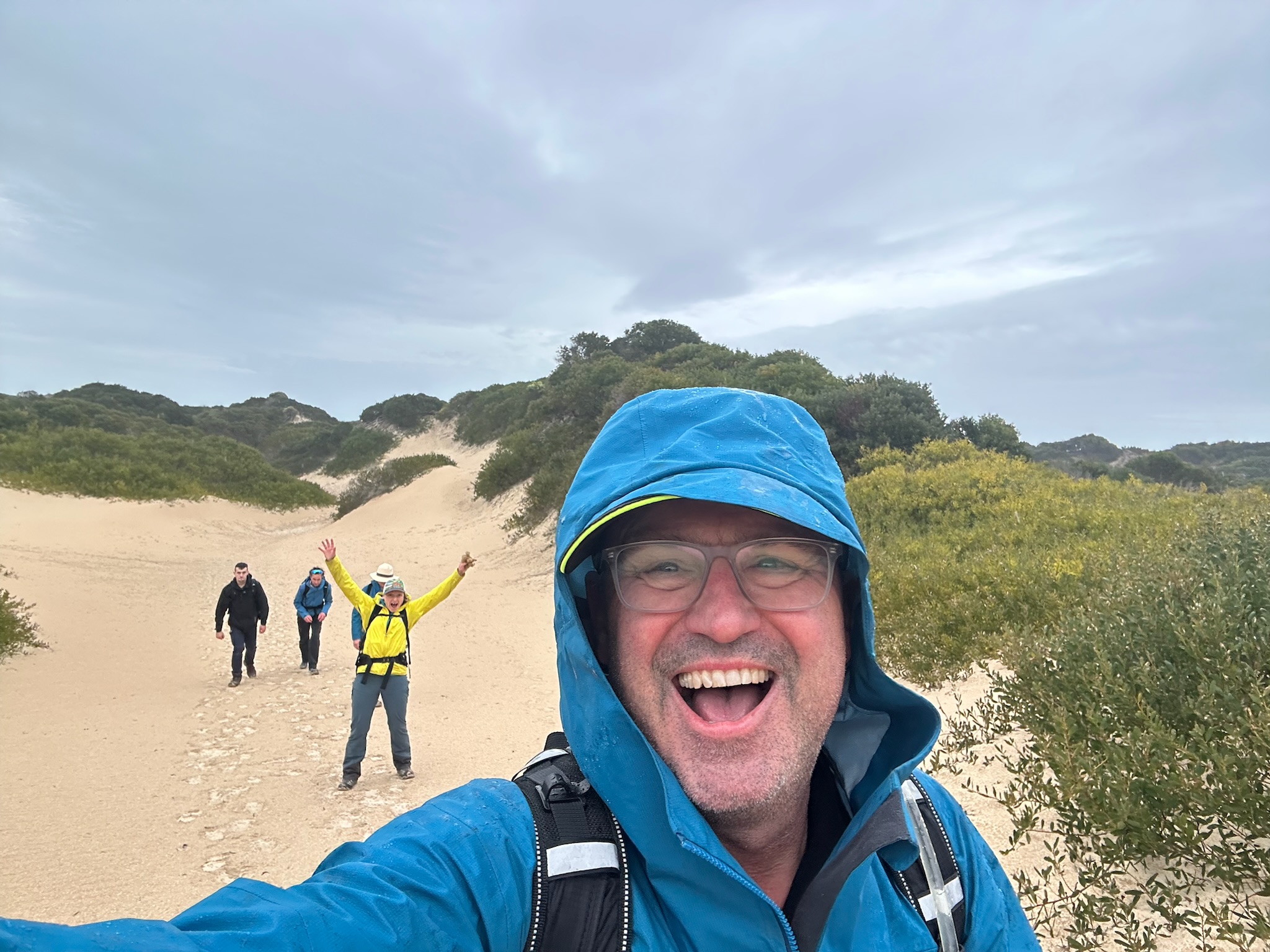 Smiling man in blue rain jacket and hood taking a selfie with three hikers in jackets walking behind him on sandy dunes with greenery under a cloudy sky.