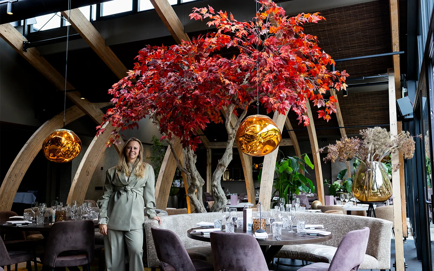 Vrouw in lichtgroten pak staat in modern restaurant met ronde tafels, paarse stoelen en grote boom met rode bladeren.