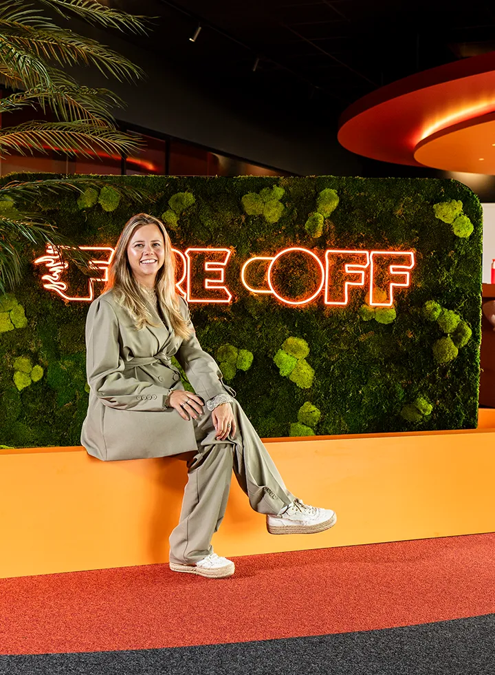 Vrouw met beige pakken en witte sneakers zittend op een oranje bank voor een groene mosmuur met neonbord 'FIRECOFF'.