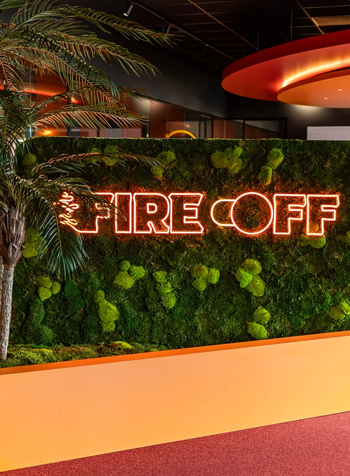 Neon teken met de tekst 'FIRE OFF' op een groene mosmuur naast een palmboom in een moderne ruimte.