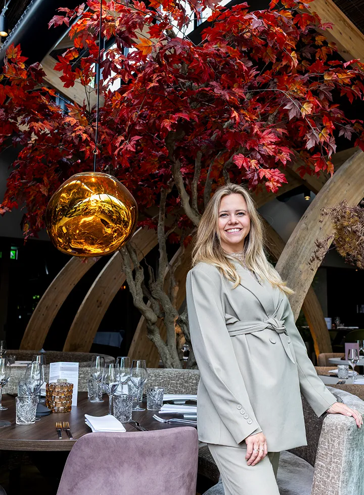 Vrouw in beige pak glimlachend in een restaurant met rood herfstblad aan bomen en een gouden hanglamp.
