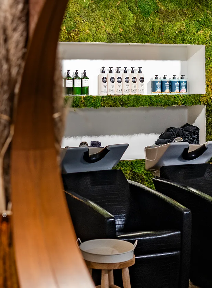 Zwarte stoelen en wasbakken voor haarwassen in een salon met een groene mosmuur en planken met verzorgingsproducten.