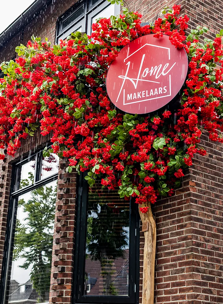 Rode bloemen rond een bord met de tekst 'Home Makelaars' aan de gevel van een bakstenen gebouw in de regen.