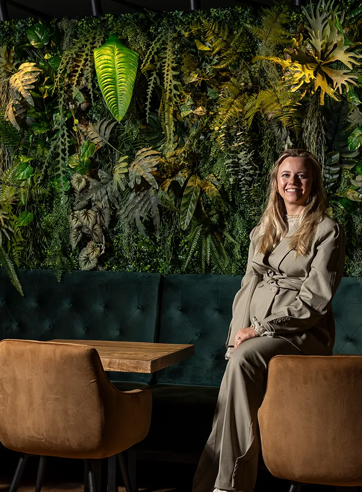 Vrouw in beige pak zittend op een bruine stoel bij een houten tafel, met een groene plantenwand op de achtergrond.