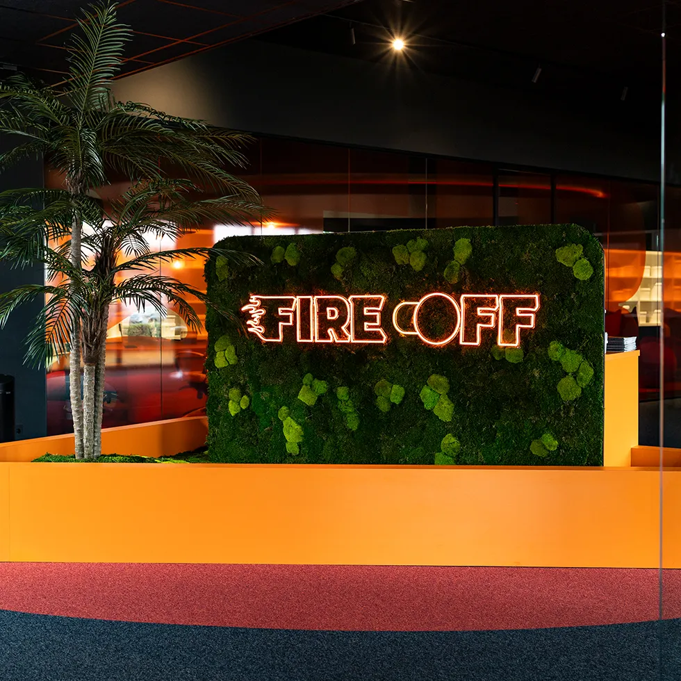 Neonlicht bord met de tekst 'FIRE OFF' bevestigd aan een groene mosmuur naast een palmboom in een modern interieur.