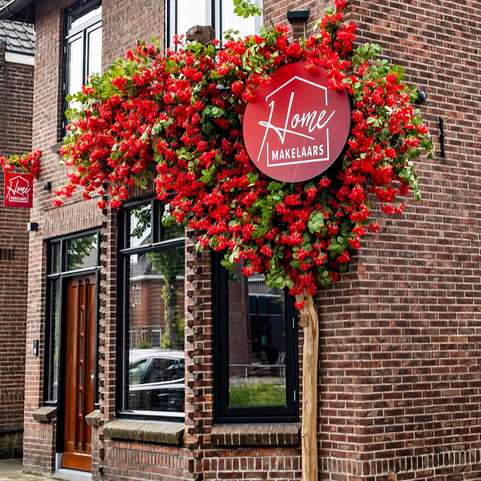 Voorkant van een bakstenen gebouw met een houten deur en een raam, versierd met een boom met rode bloemen en een rood bord met de tekst 'Home Makelaars'.