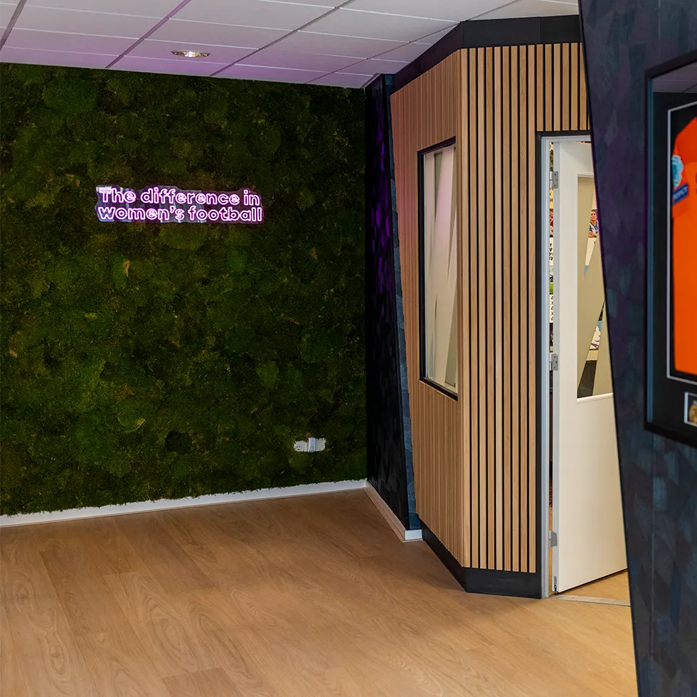 Een moderne kamer met een groene mosmuur en een neonbord met de tekst 'The difference in women's football', naast een witte deur en houten wandpanelen.