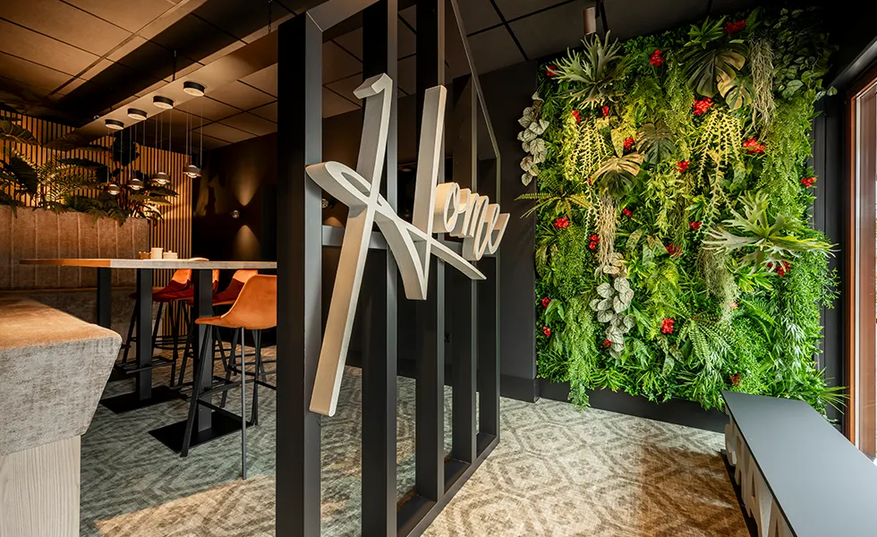 Moderne interieur met groene moswand, houten tafel met hoge stoelen en decoratief 'Home' bord aan zwart metalen frame.