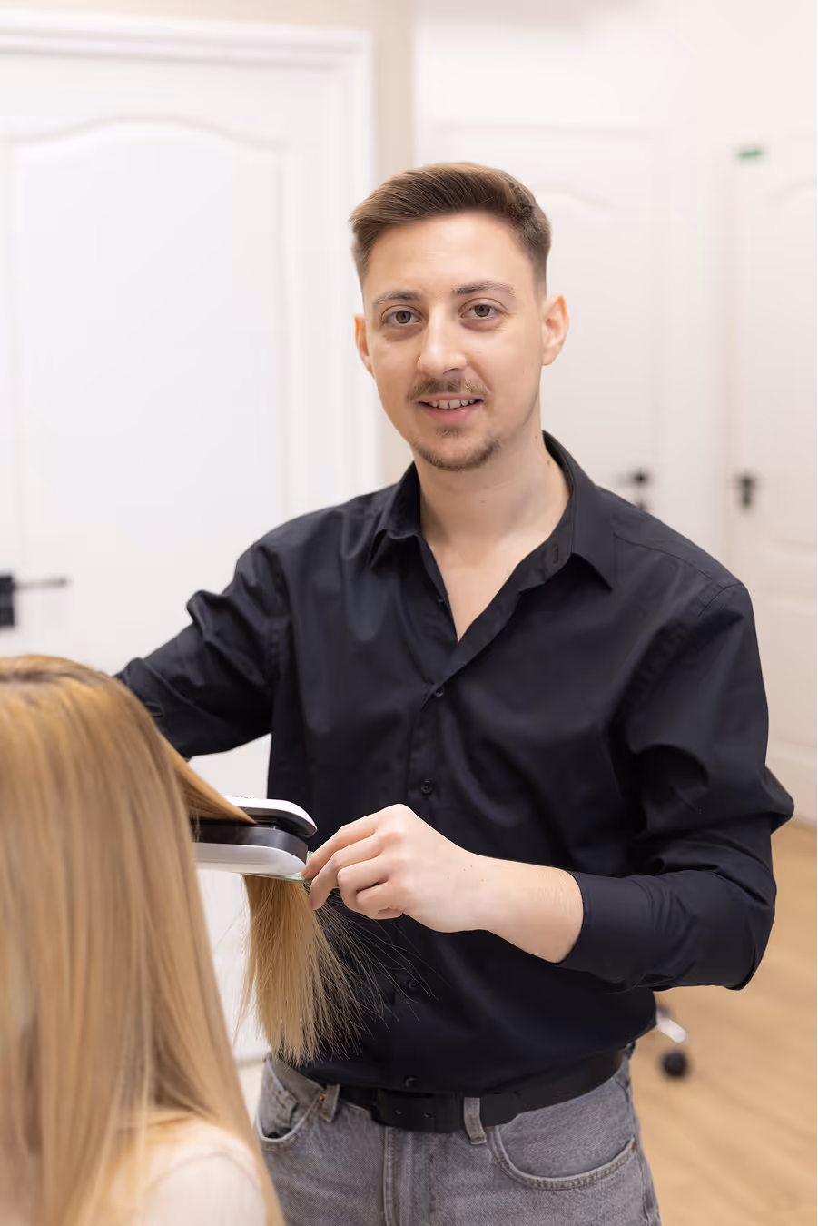 Férfi fodrász egy szalonban egy székben ülő nő hosszú szőke haját vasalja.