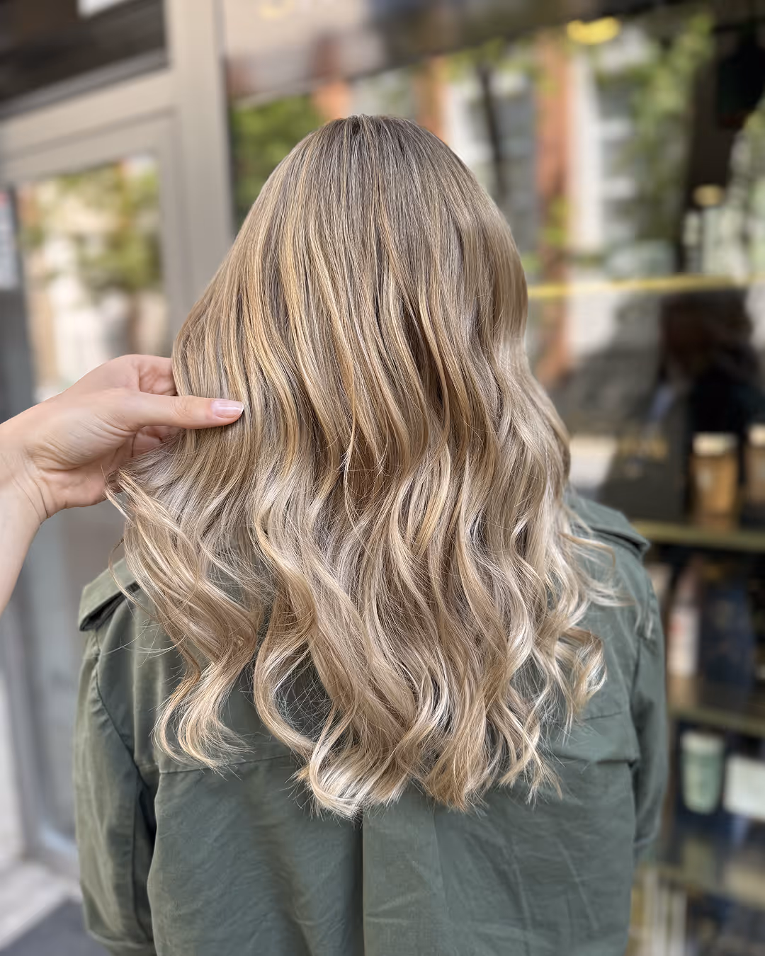 Egy hosszú, hullámos, finoman melírozott szőke hajú személy hátulnézete, amelynek egyik oldalát egy kéz tartja.