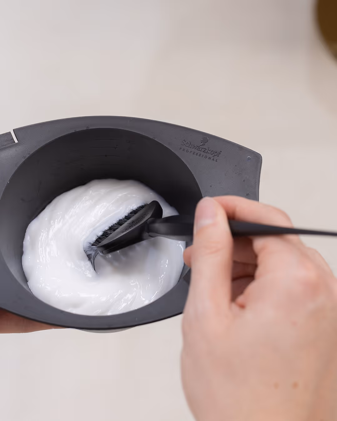 Kéz egy fekete keverőtálat tart, benne fehér hajfestéket kever egy ecsetes applikátorral.