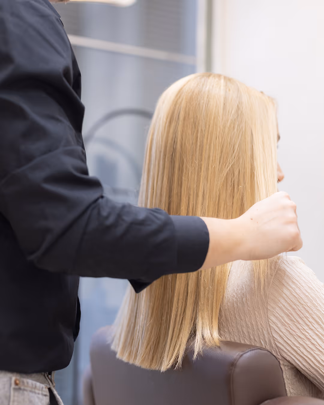 Fodrász egy szalon székben ülő nő egyenes szőke haját formázza.