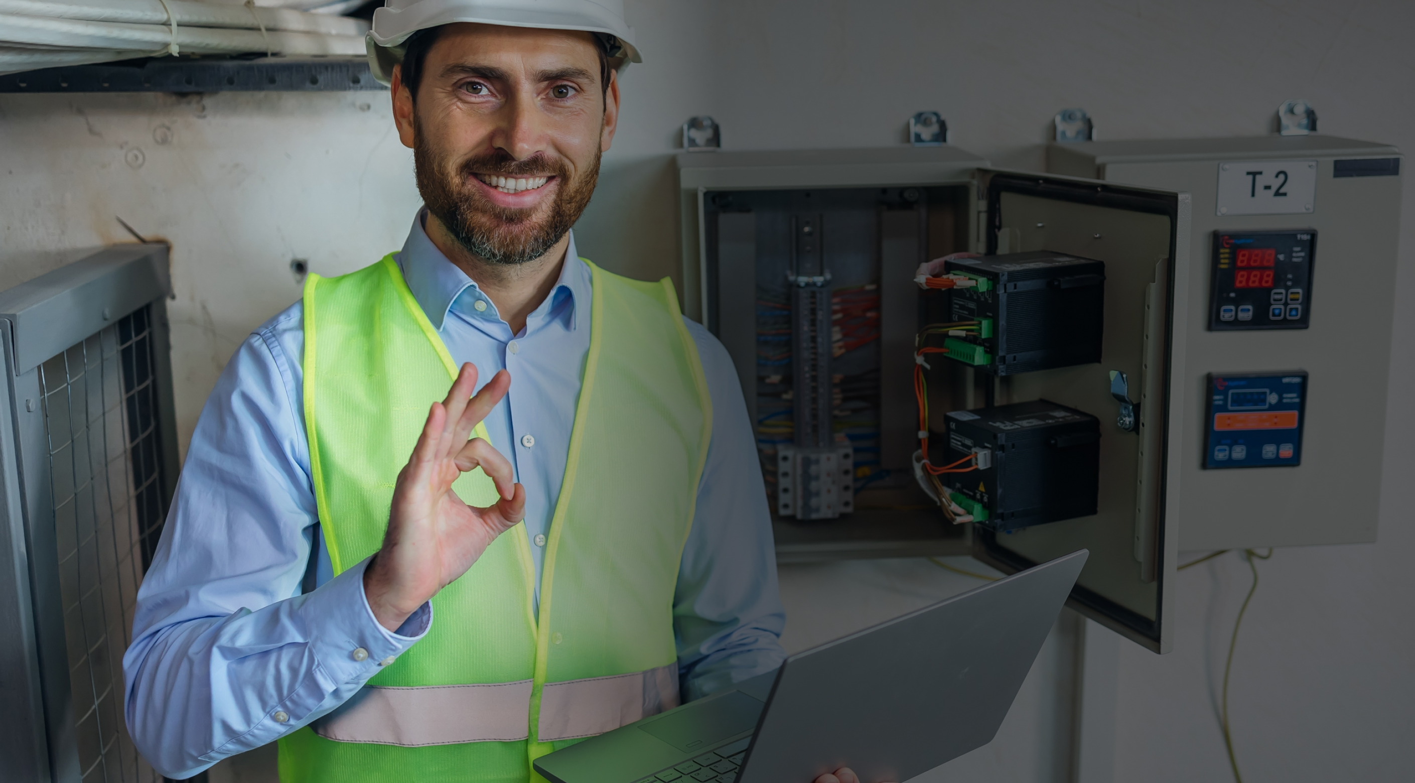 Smiling technician in a hard hat and safety vest beside an electrical panel