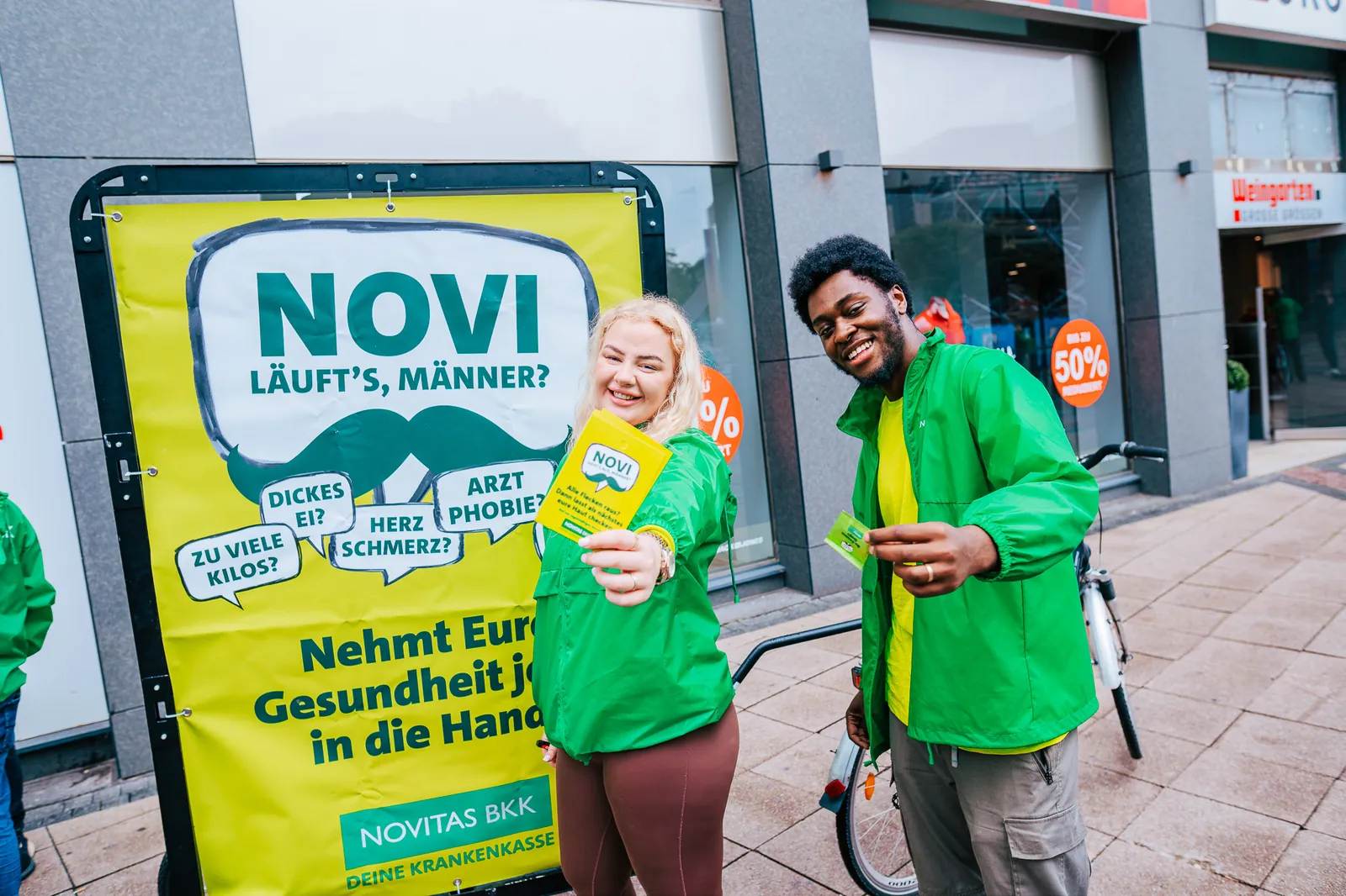 Zwei Promoter in grünen Jacken verteilen Flyer vor einem Kampagnenplakat