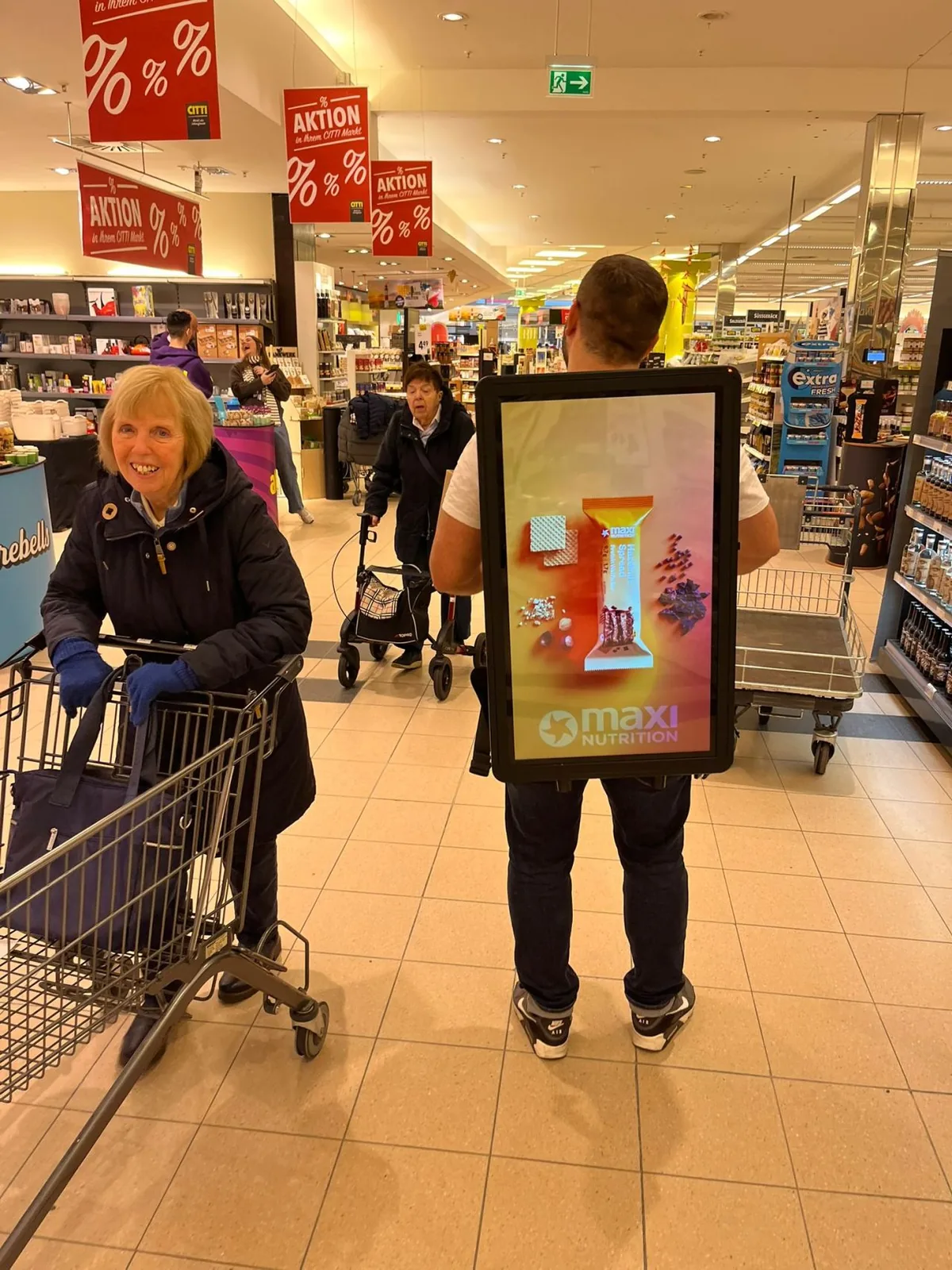 Walking Screen im Drogeriemarkt neben einer Kundin mit Einkaufswagen
