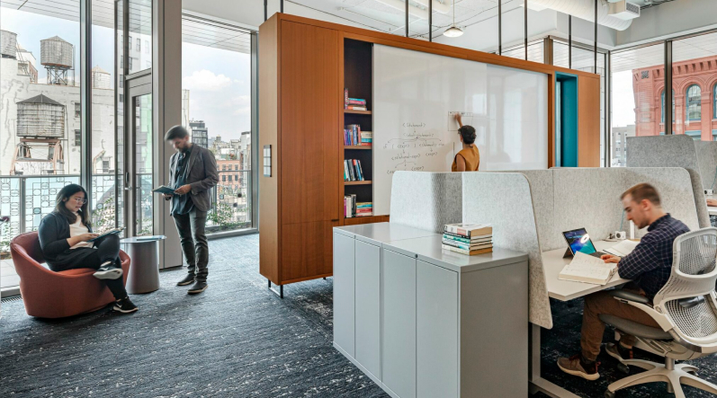 Modern office with three people: one woman sitting on a pink chair reading, one man standing reading a notebook near large windows, and one person writing on a whiteboard behind a wooden bookshelf.