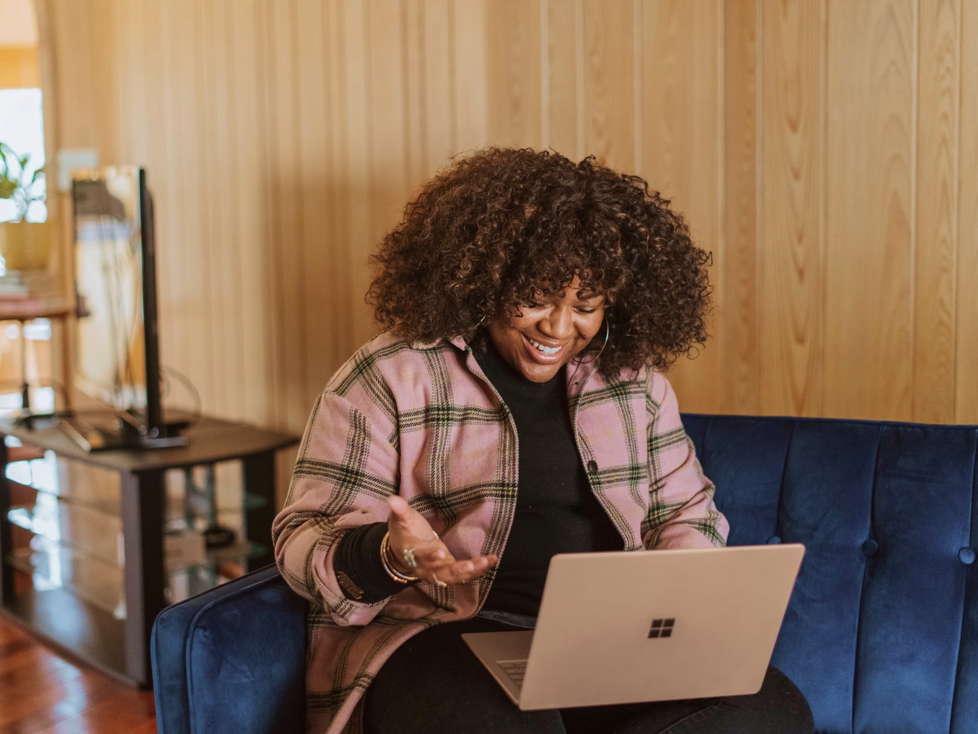 Woman with curly hair smiling and gesturing while on a video call in a wooden-paneled room.