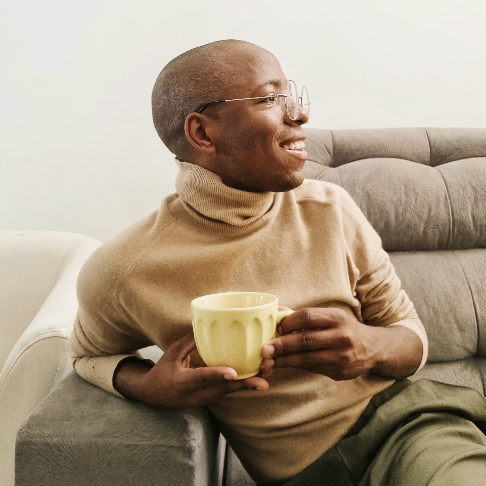 Smiling person in glasses and beige turtleneck sitting on a grey sofa, holding a yellow mug.