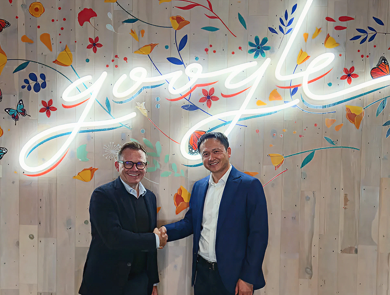 Two men in suits shaking hands in front of a wall with a neon Google logo and colorful floral decorations.