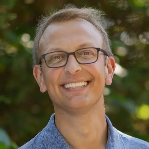 Smiling man with glasses in a green outdoor setting.