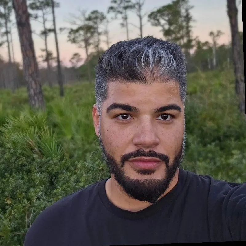 Man with a beard and short gray hair smiling in a forested area.