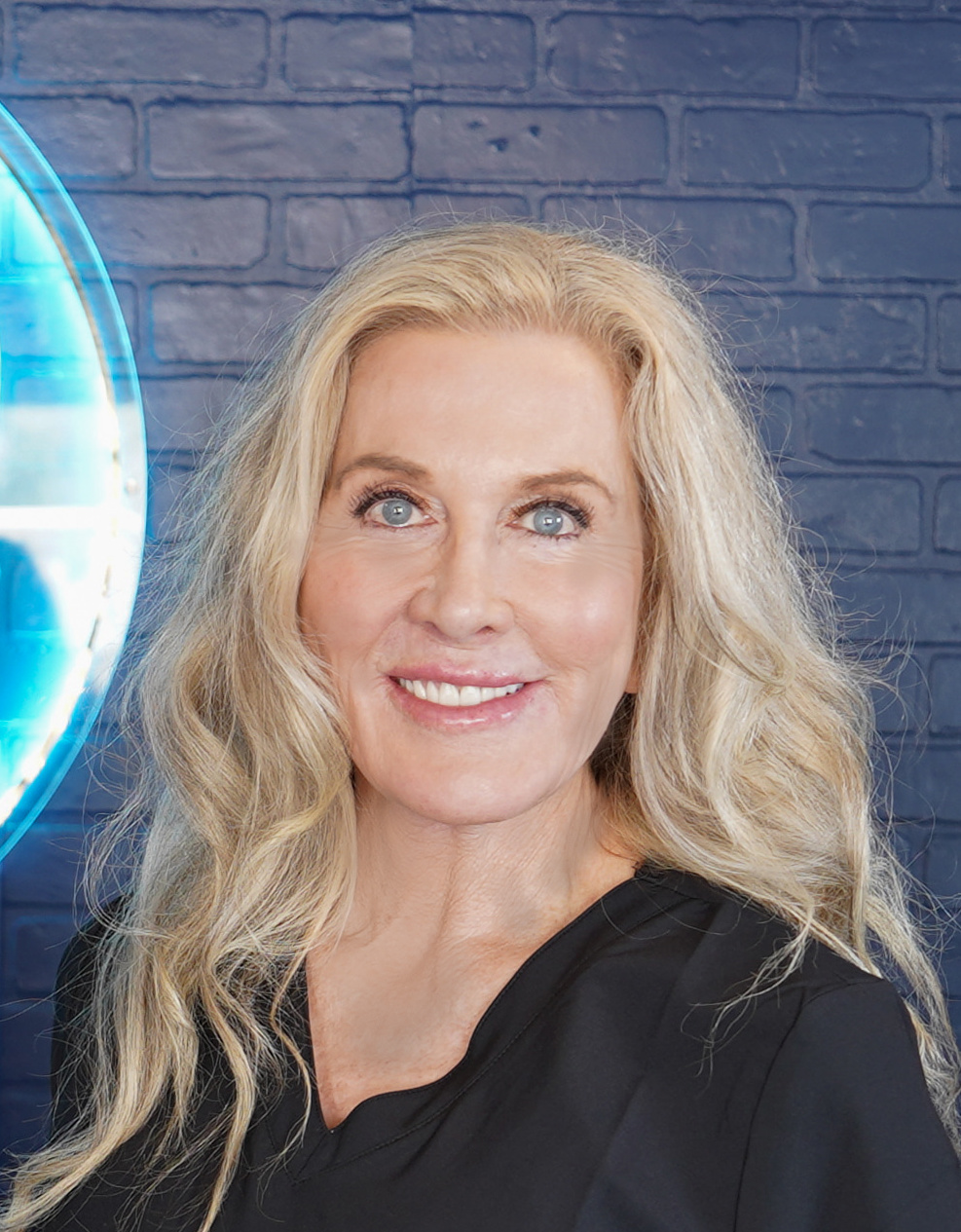 Middle-aged woman with shoulder-length blond hair wearing a black jacket and pearl necklace, smiling in front of a facial rejuvenation poster.