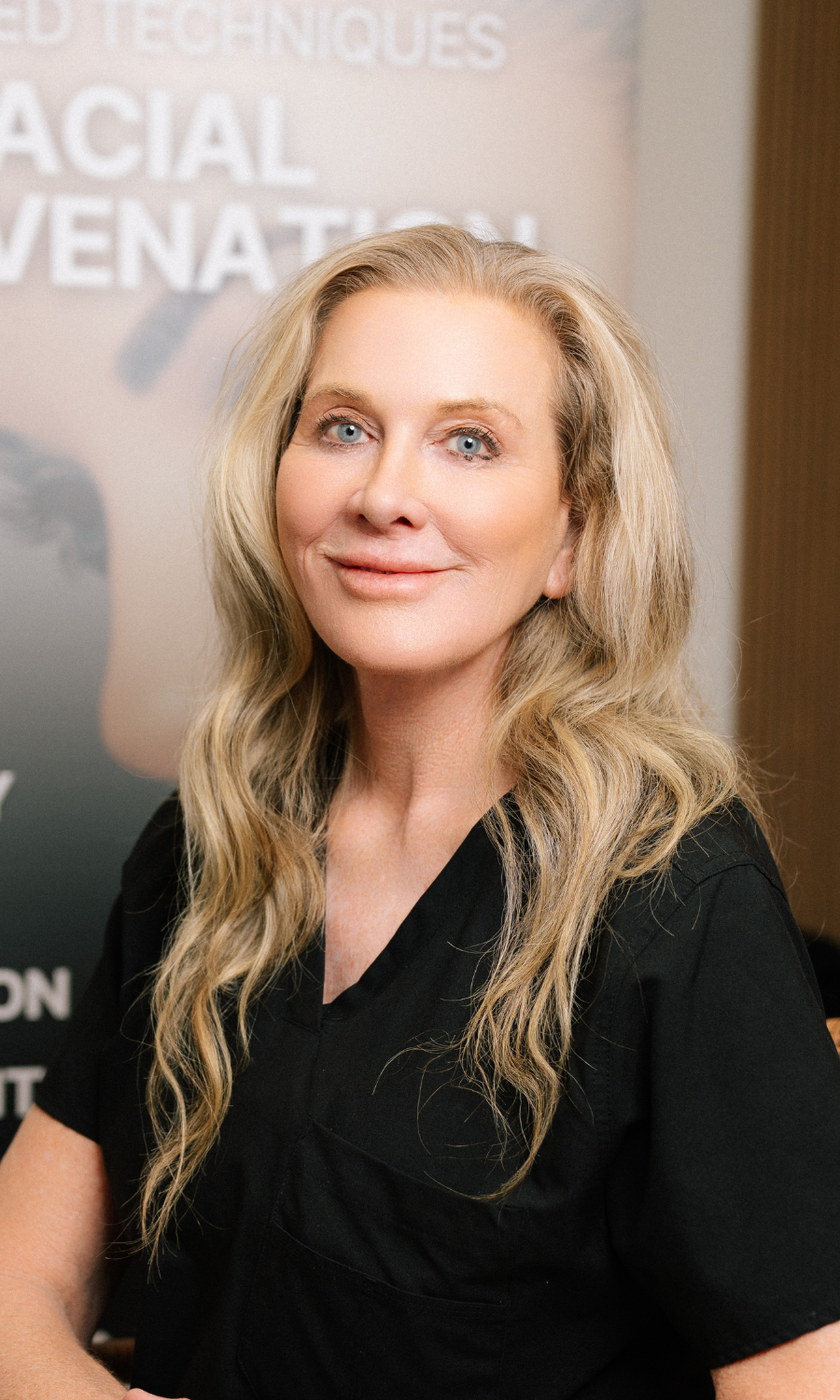 Middle-aged woman with shoulder-length blond hair wearing a black jacket and pearl necklace, smiling in front of a facial rejuvenation poster.