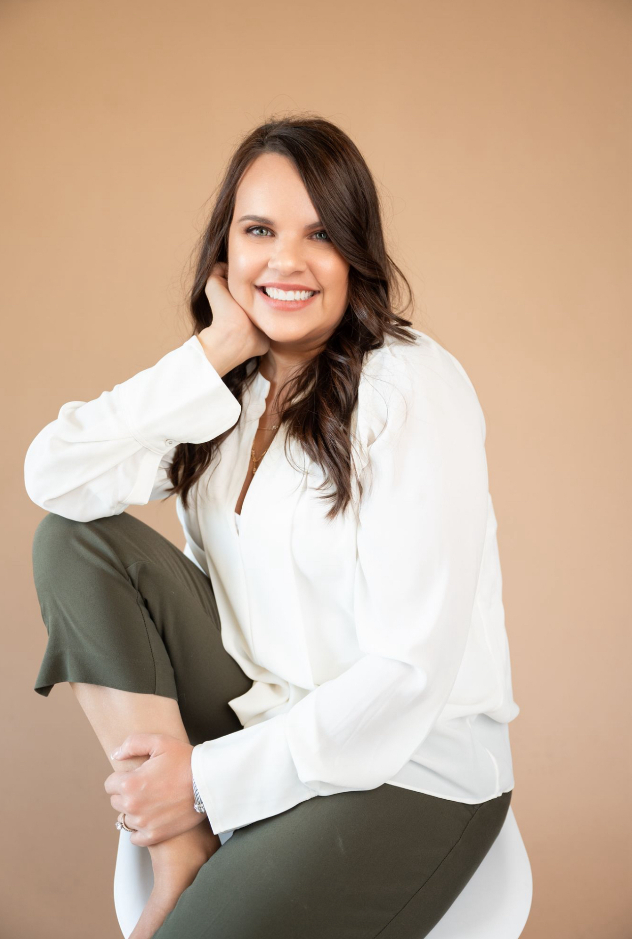 Middle-aged woman with shoulder-length blond hair wearing a black jacket and pearl necklace, smiling in front of a facial rejuvenation poster.