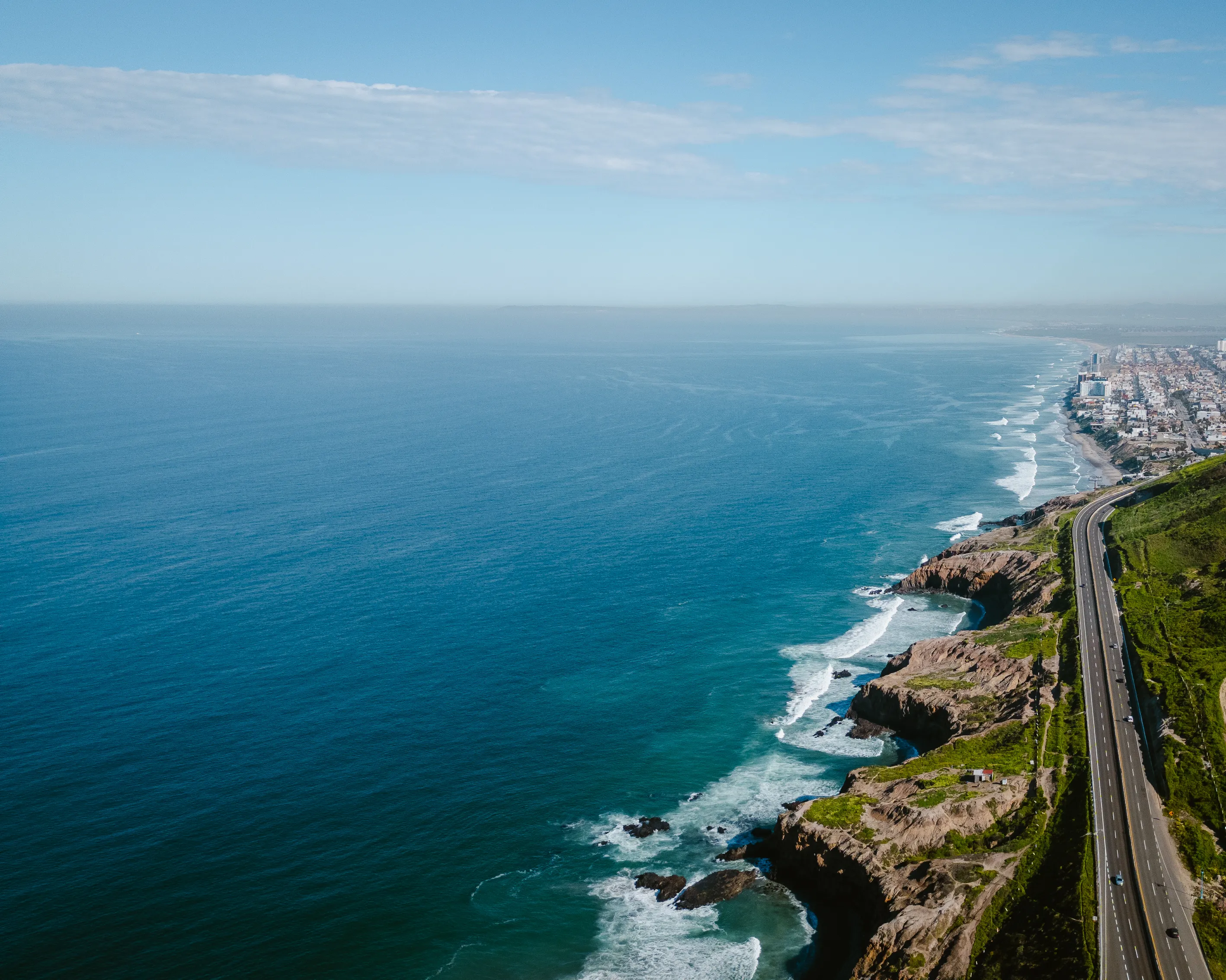 Vista aérea de una carretera costera junto al océano con acantilados rocosos y una ciudad al fondo.