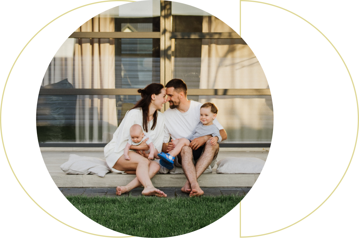 A happy family of four sitting on a porch step with green grass in front, parents smiling at each other, mother holding a baby, and father holding a toddler.